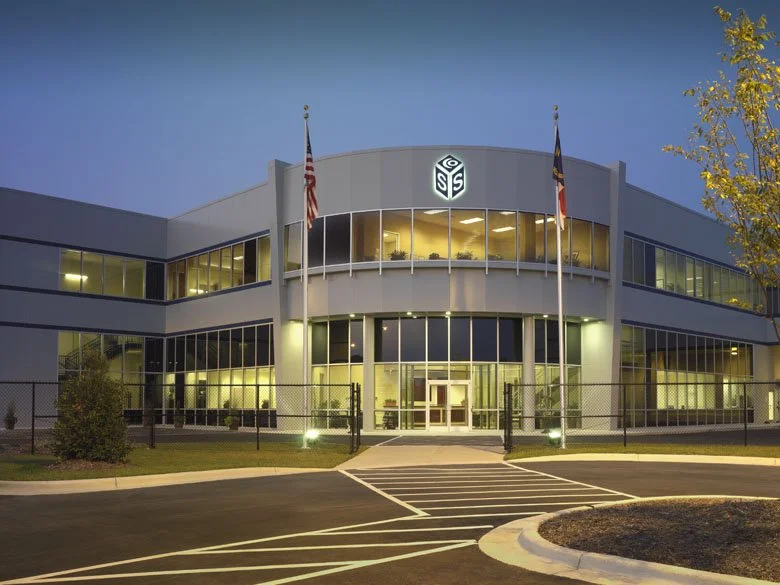 Exterior view of a modern office building with glass windows, illuminated from within, showing the company's logo on the front. Two flags are displayed in front of the building, and the area is surrounded by a parking lot with marked spaces.