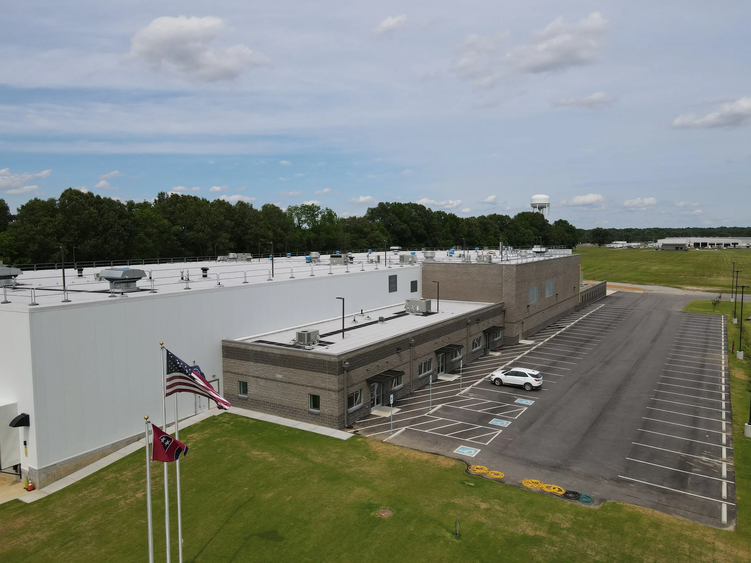 An aerial view of a large commercial building with a parking lot, some parking spaces marked for handicapped, and flags flying outside. The building has a flat roof with various HVAC units and a water tower in the background.