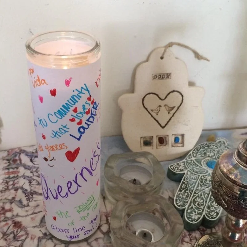 A light tall glass jar prayer candle decorated with words and images as an homage to the brilliance of queer latinx vibrancy sitting to next to unlit shabbat candles, decorative hamsas, and a bismam holder.