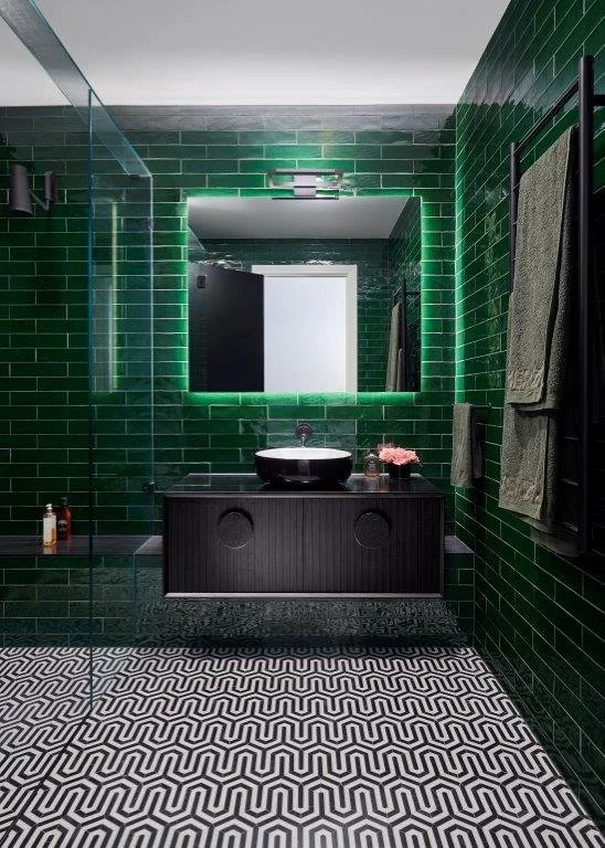 Modern bathroom with green tiled walls, large illuminated mirror, black vanity with vessel sink, black towel rack with green towels, and black and white geometric patterned floor.