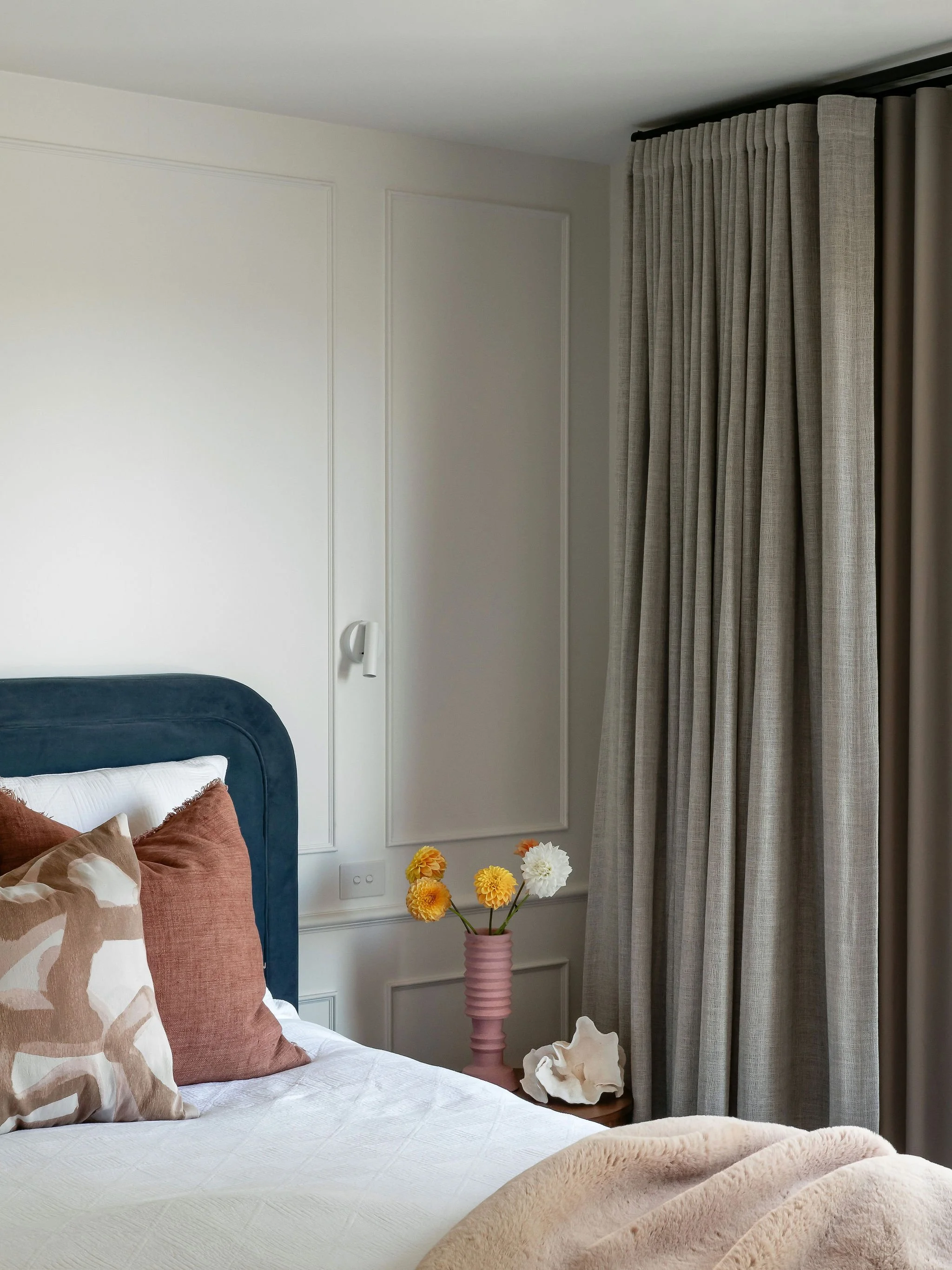 A bedroom corner with a bed, decorative pillows, a side table with a pink vase holding flowers, a shell decor piece, and curtains.