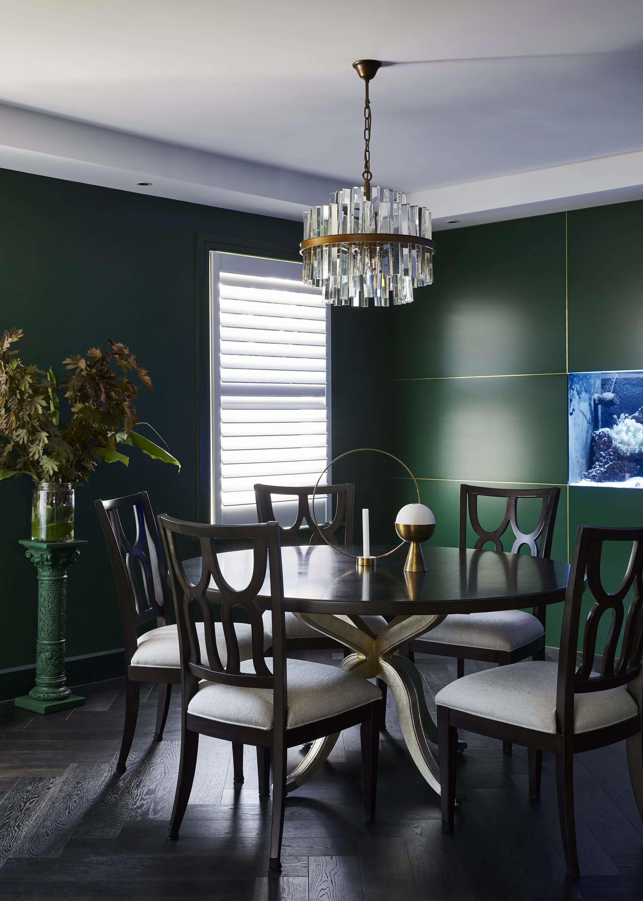 Dining room with a dark wood round table, six matching chairs with beige cushions, and a modern chandelier with hanging glass pieces. Green accent wall with a window featuring white shutters, a tall green plant on a decorative green pedestal, and a b