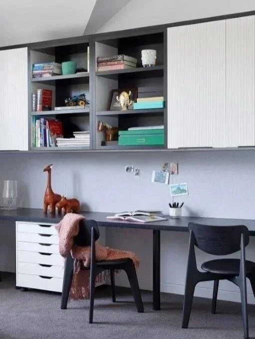 A home office with a black desk, two black chairs, and upper gray shelving filled with books, plants, and decorative items. There are toys like a giraffe and a horse on the desk, along with a pink blanket, and photos and notes pinned on the wall behi