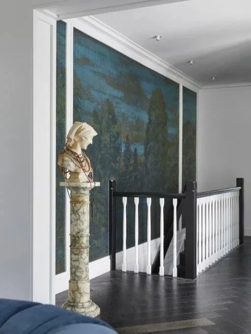 Interior of a house or gallery with a staircase, decorated with a classical bust on a pedestal and a large landscape painting on the wall.