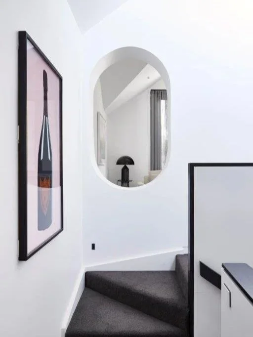 Interior view of a modern home with white walls, a round mirror, a framed artwork of a wine bottle, a black table lamp, and a staircase with dark carpeting.
