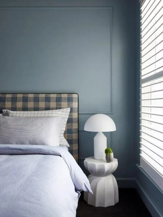 A bedroom corner with a bed, checkered headboard, white pillow, and a bedside table with a white lamp and small plant, next to window with white blinds.