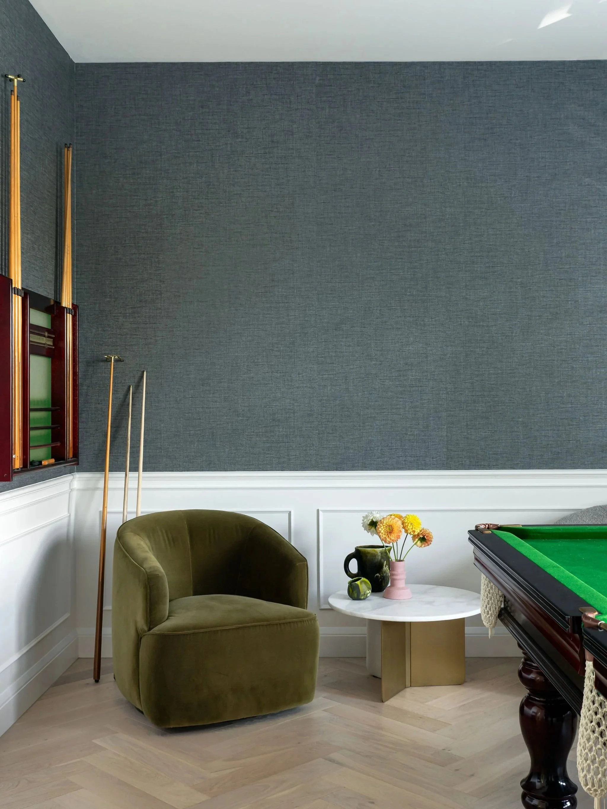 Corner of a room with a green velvet armchair, a small white round table with a black and pink vase holding flowers, a pool table with green felt, and wall-mounted pool cues against dark textured wallpaper.