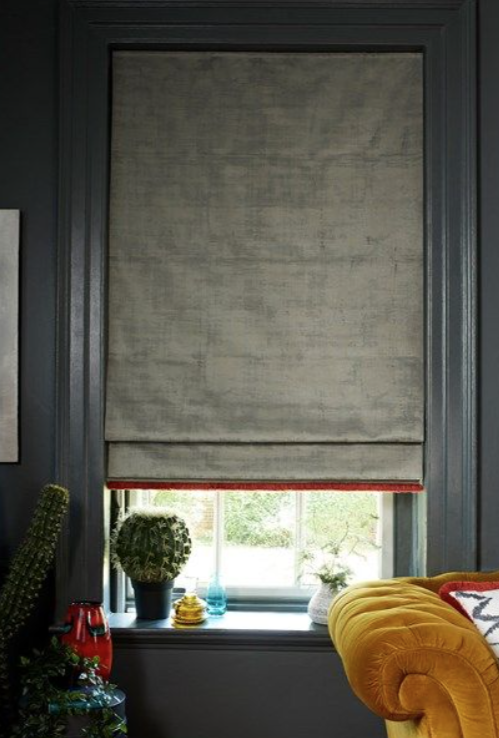 Modern living room with large windows covered by floral-patterned blinds, a black pendant lamp hanging from the ceiling, a floor lamp with a beige shade, and a white sofa with a blue pillow. Greenery is visible outside the window.