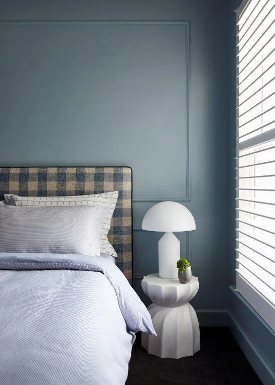 A bedroom with a light blue wall, a checkered bed headboard, white bed linens, a bedside table, a white lamp, and a small plant in a gray pot near a window with white blinds.