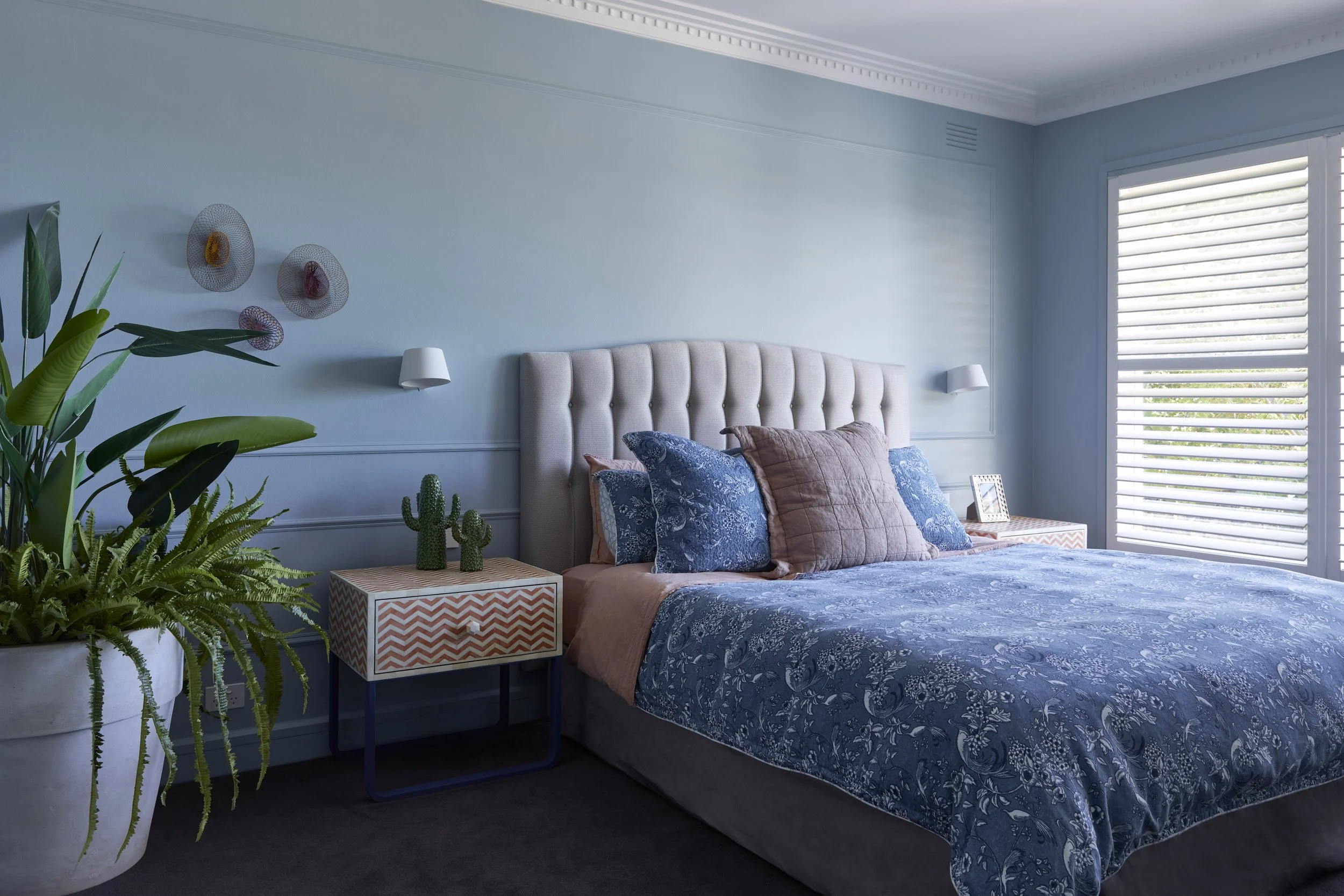 Bedroom with light blue walls, white upholstered headboard, blue and peach bedding, plants, and window with blinds.