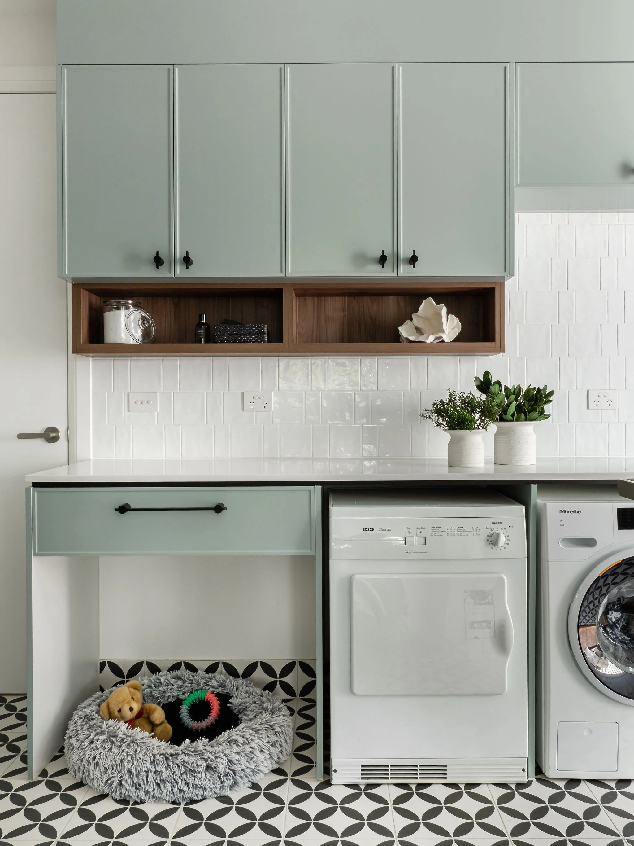 Laundry room with pastel green cabinets, white tiled wall, a washing machine, and a pet bed with toys on black and white patterned floor.