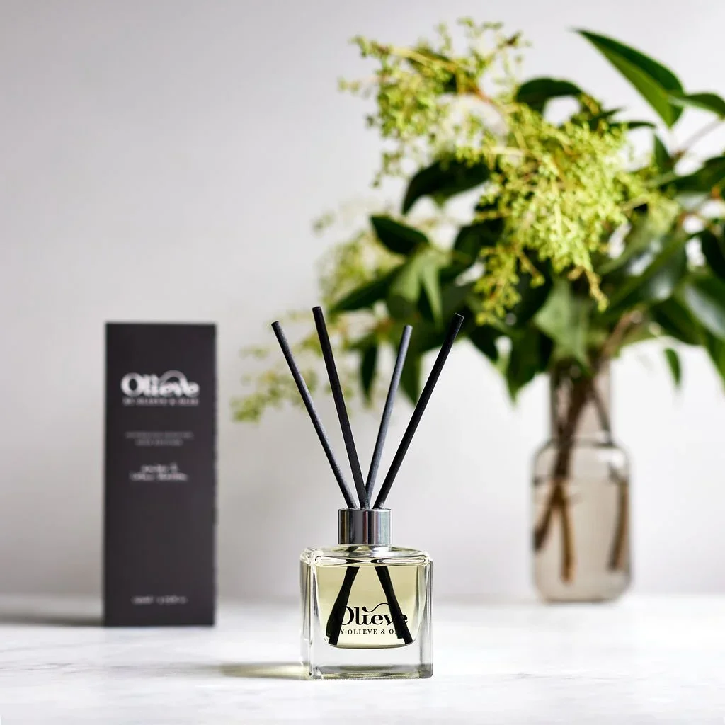 A glass reed diffuser with black reeds and a label that reads "Olea" in front of a potted plant with green leaves and cream-colored flowers, and a black box with white text in the background.