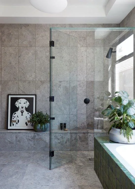 Modern bathroom with gray tiled walls and floor, a glass shower door, a black showerhead, a framed dog artwork, green plants on a tiled ledge near a bathtub, and a window.