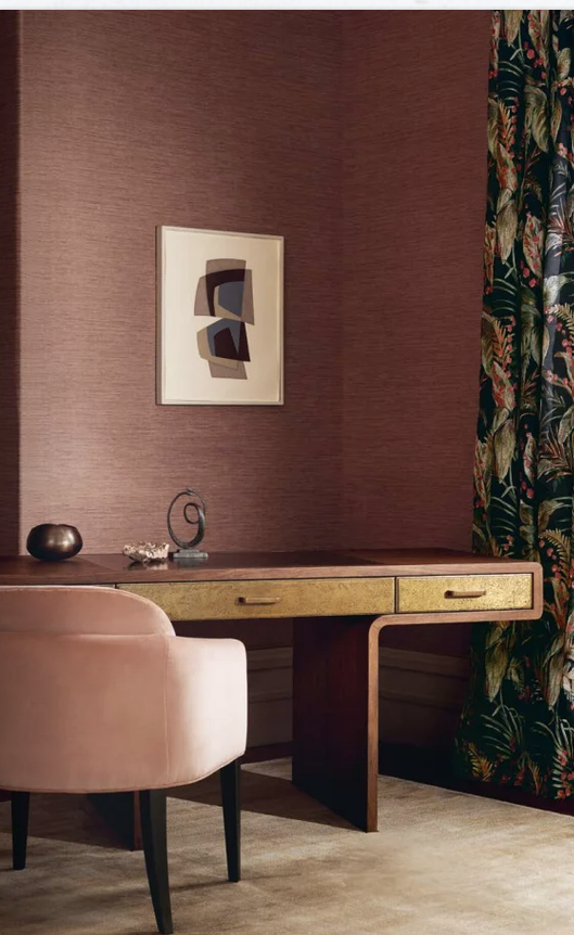 A corner of a room featuring a wooden desk with two drawers, a pink upholstered chair, a small decorative bowl, and a modern abstract art piece hanging on a textured pink wall. Dark floral curtains are partially visible on the right side.