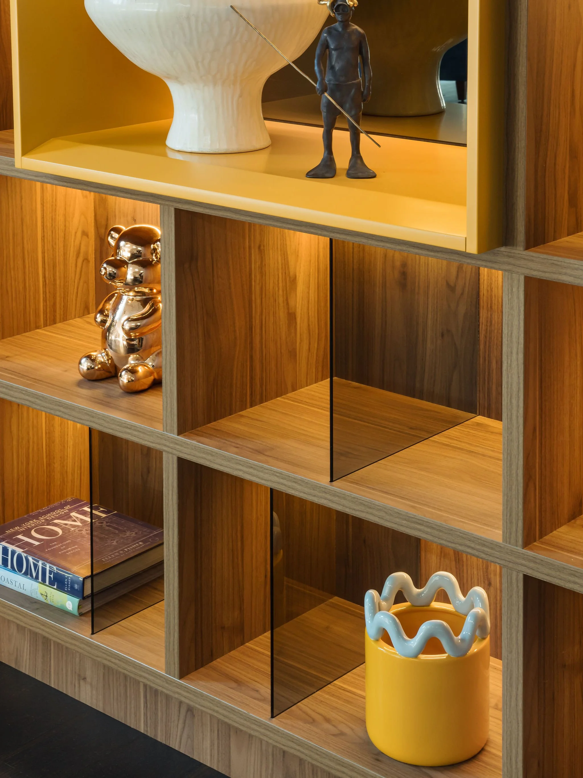 A wooden shelving unit with navigational glass panels separating different sections. The shelves contain decorative objects including a white vase, a bronze statue of a boy with a fishing rod, a shiny gold teddy bear sculpture, a few books, and a bri