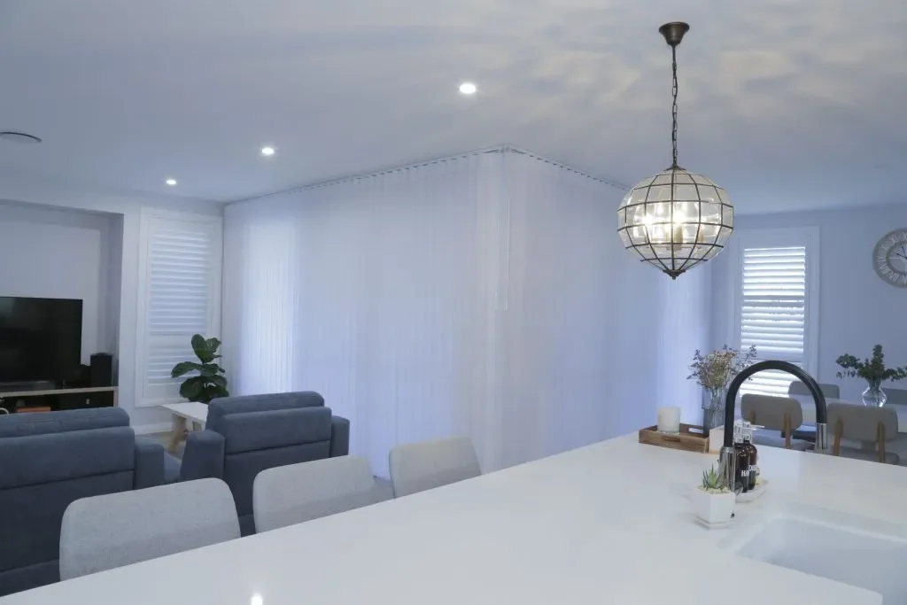 Bright open-concept living room and kitchen with white walls, a large island, dining area, and modern seating, illuminated by a hanging pendant light.