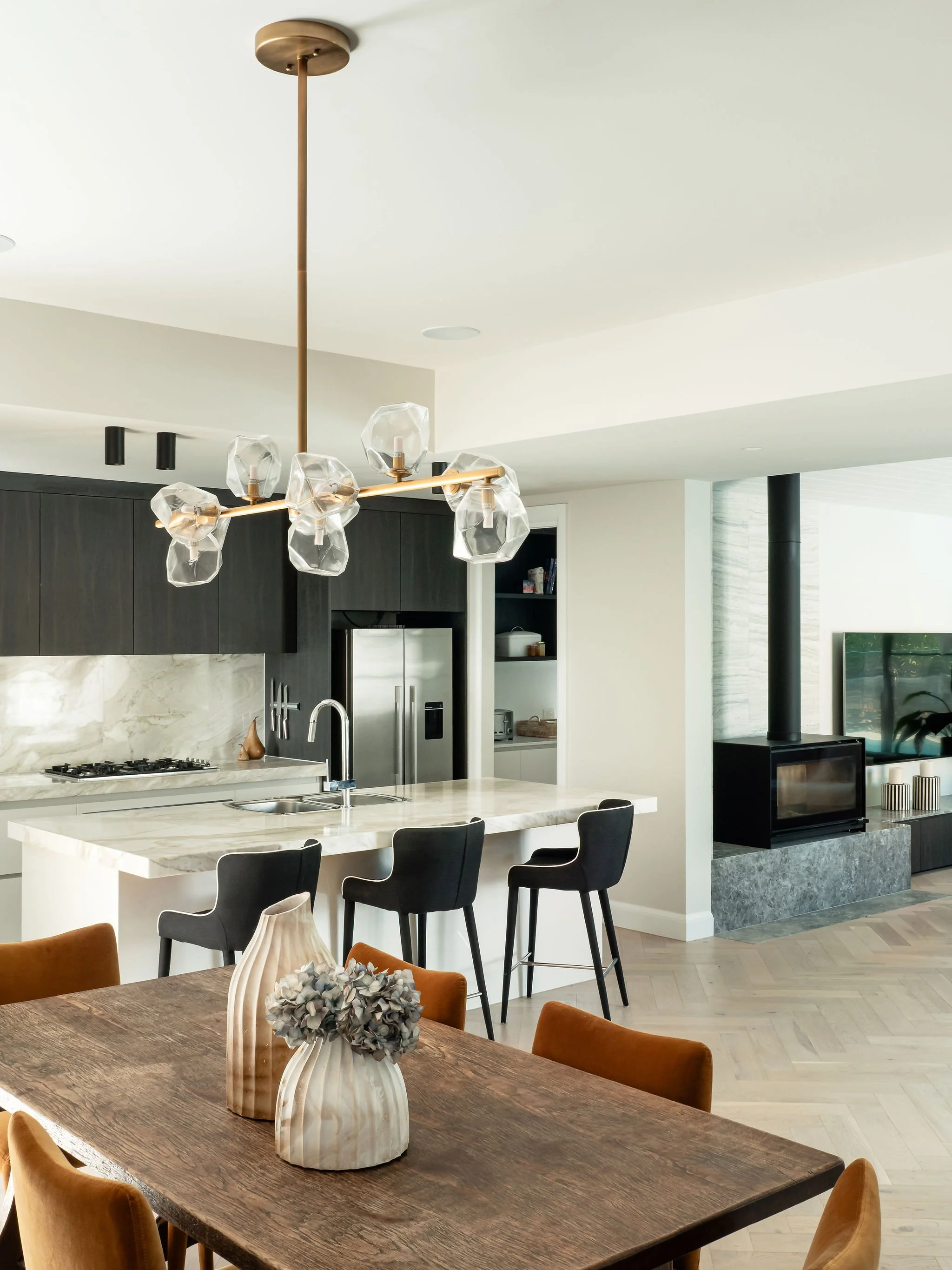 Open-concept kitchen and dining area with a wooden dining table, orange chairs, vases with flowers, a marble kitchen island with black chairs, a modern chandelier, and a living space with a wood stove and TV.