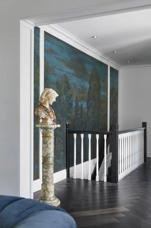 Interior view of a staircase landing with a decorative bust on a pedestal, a large landscape painting on the wall, white walls, and dark hardwood floors.