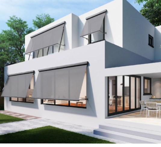 Modern white house with large windows and gray awnings, outdoor patio with dining table, surrounded by greenery.