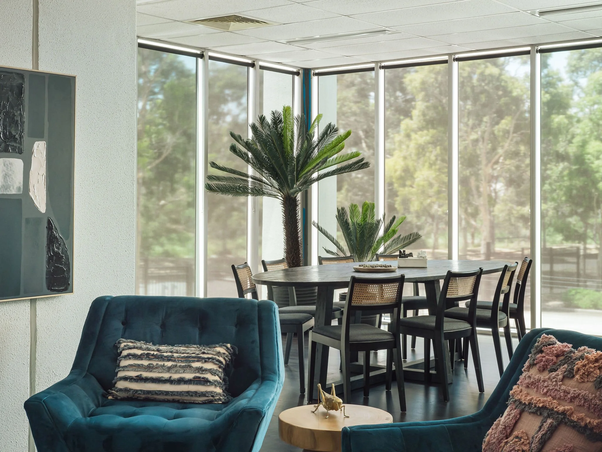 Interior of a modern living and dining area with large windows, two blue armchairs, a small wooden side table with a gold decorative horse, a round dining table with eight chairs, and two large potted plants in the background.