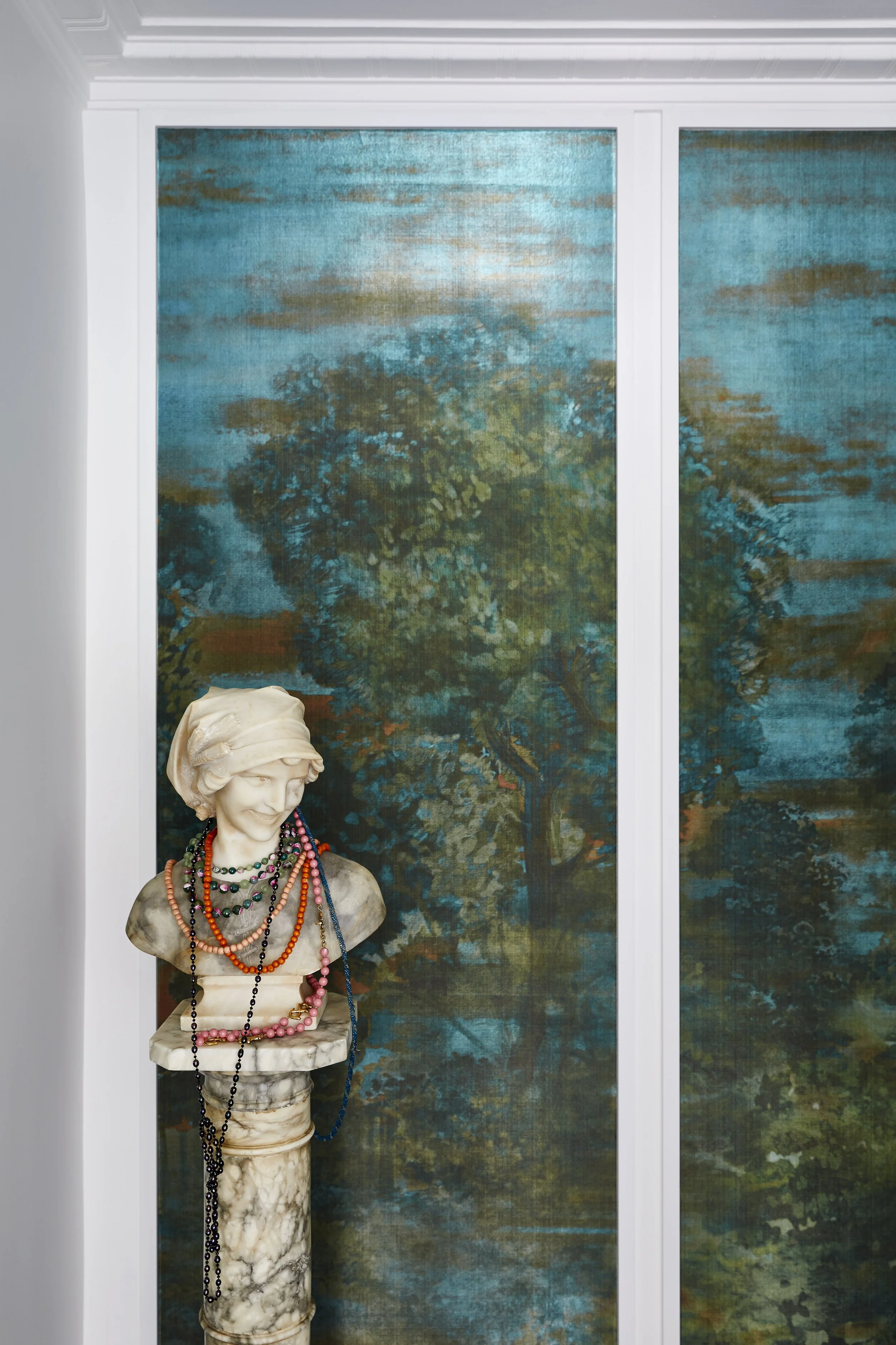 Bust of a woman wearing multiple beaded necklaces, with a textured headscarf, set against a landscape painting on a wall with sliding doors.