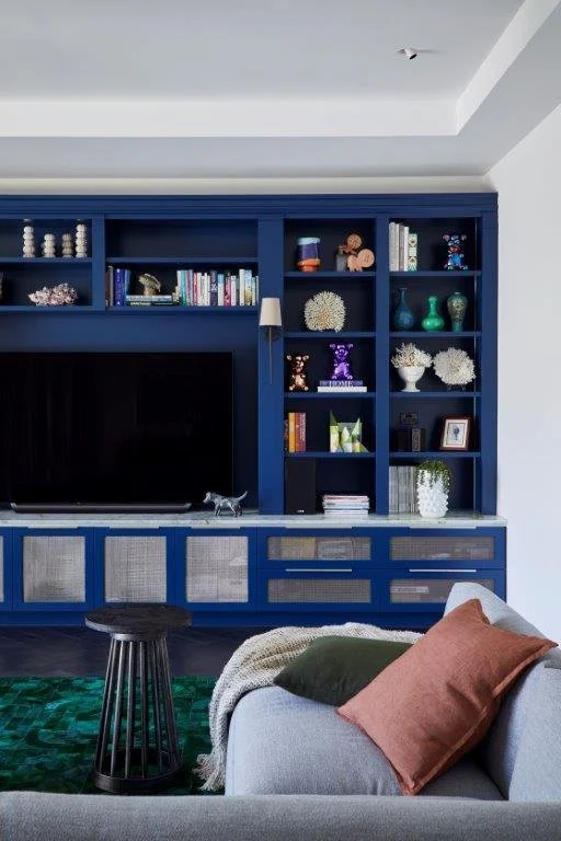 Living room with dark blue built-in shelves and cabinets, flat-screen TV, decorative items, books, and a small silver sculpture; gray sofa with brown and dark green pillows, green patterned rug, black round side table, and white walls.