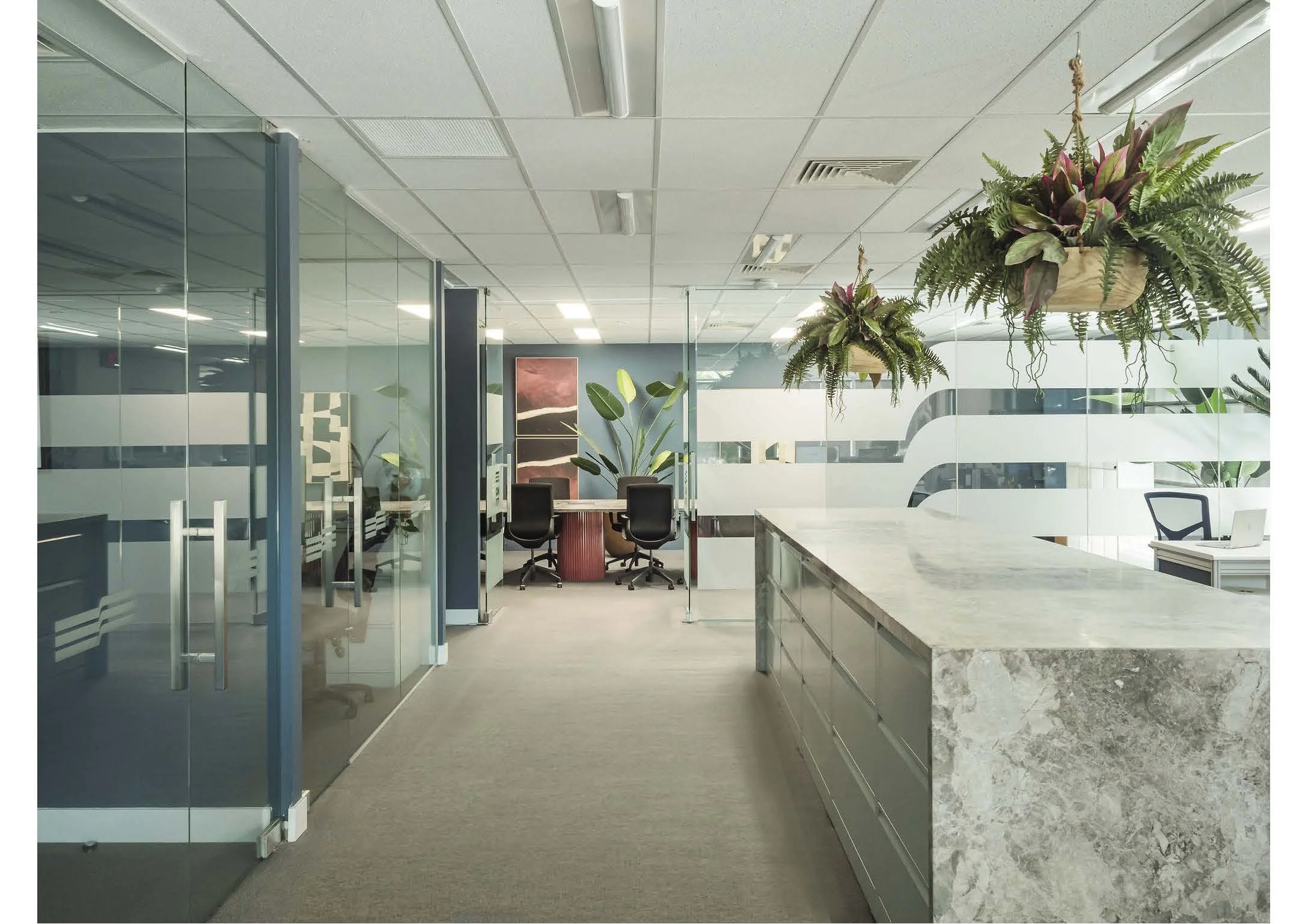 Modern office space with glass partitions, a conference room with chairs, a marble kitchen island, hanging plants, and a workspace with a laptop.