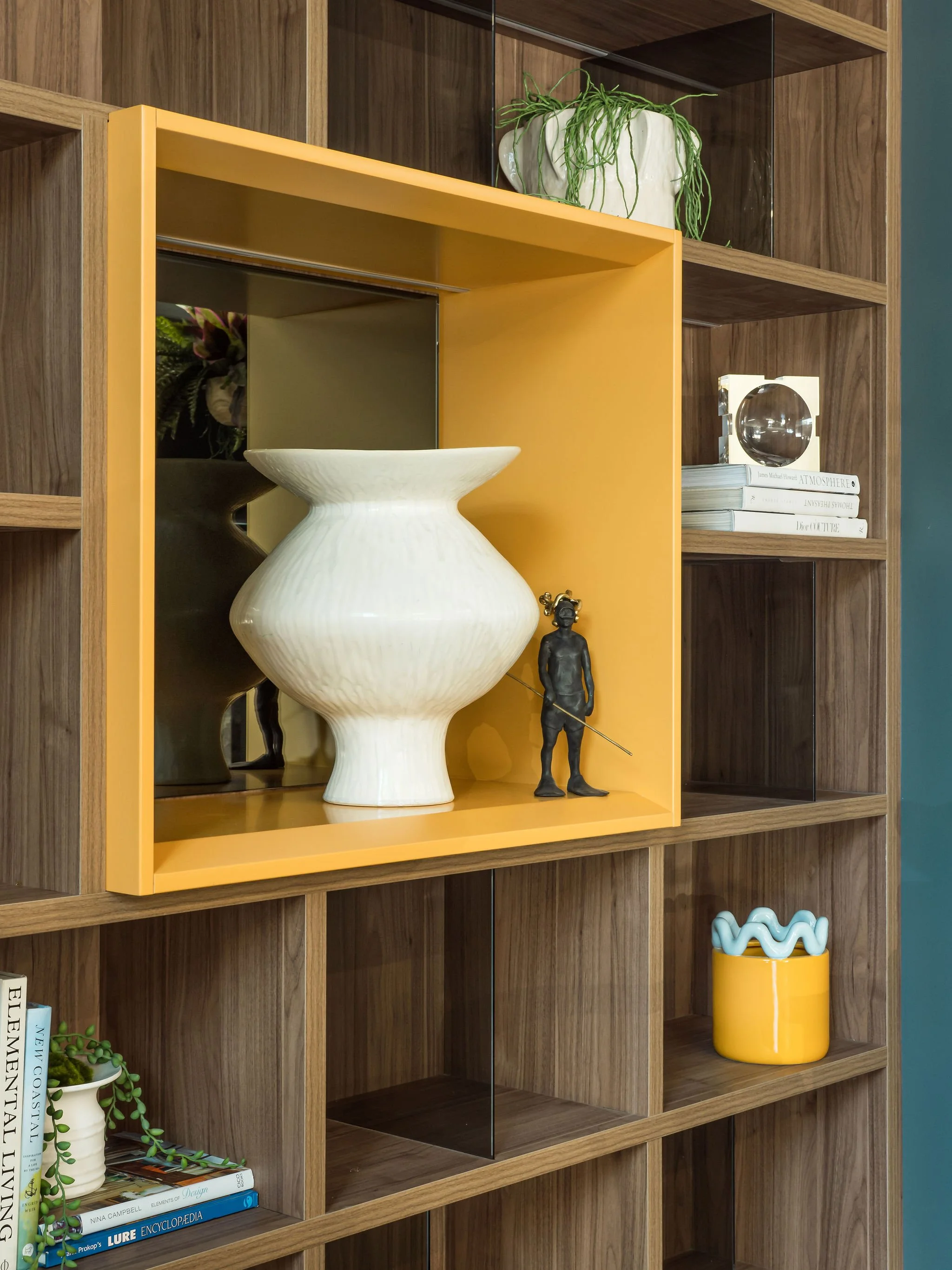 Decorative shelf with a large white ceramic vase, a small black sculpture of a person, plants, books, and a yellow container with blue decorative objects.
