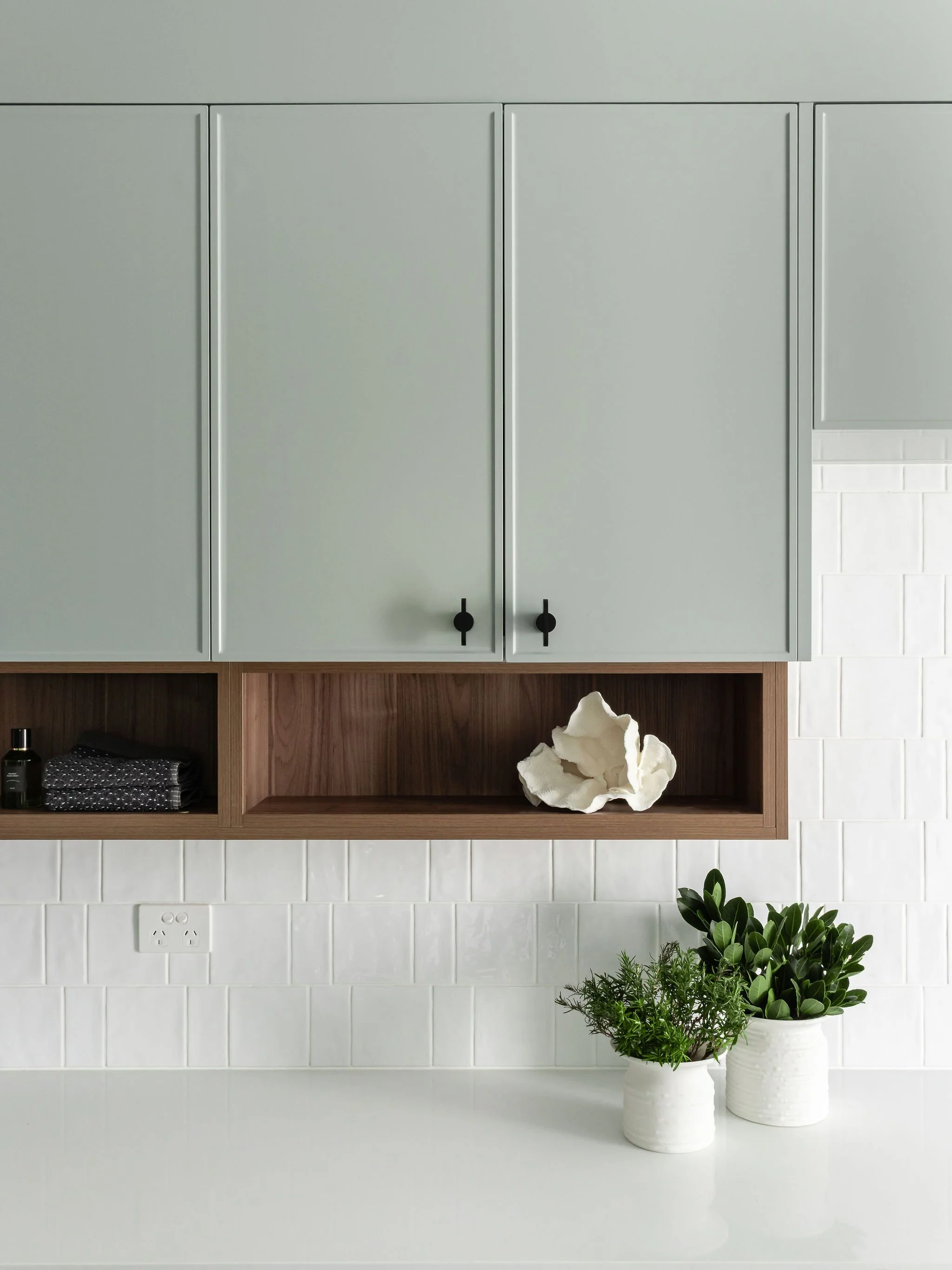 Kitchen with light green cabinets, wooden open shelf, white countertop with potted green plants, and white tile backsplash.