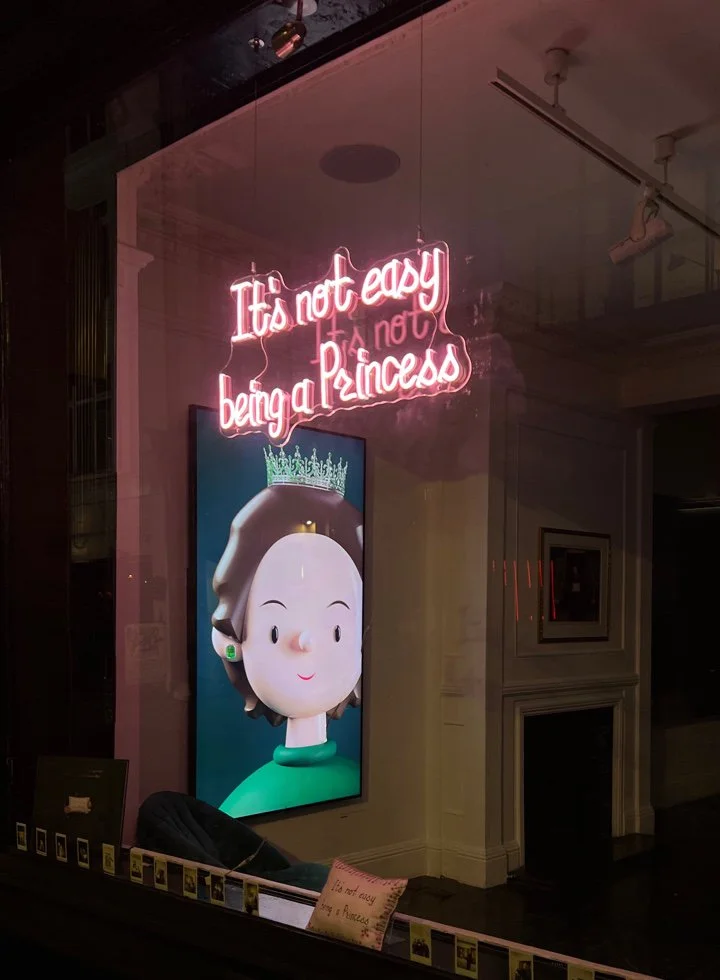 A neon sign that says 'It's not easy being a Princess' lit in pink, hanging in a window beside an animated digital portrait of a cartoon princess with a crown, earrings, and a green shirt.