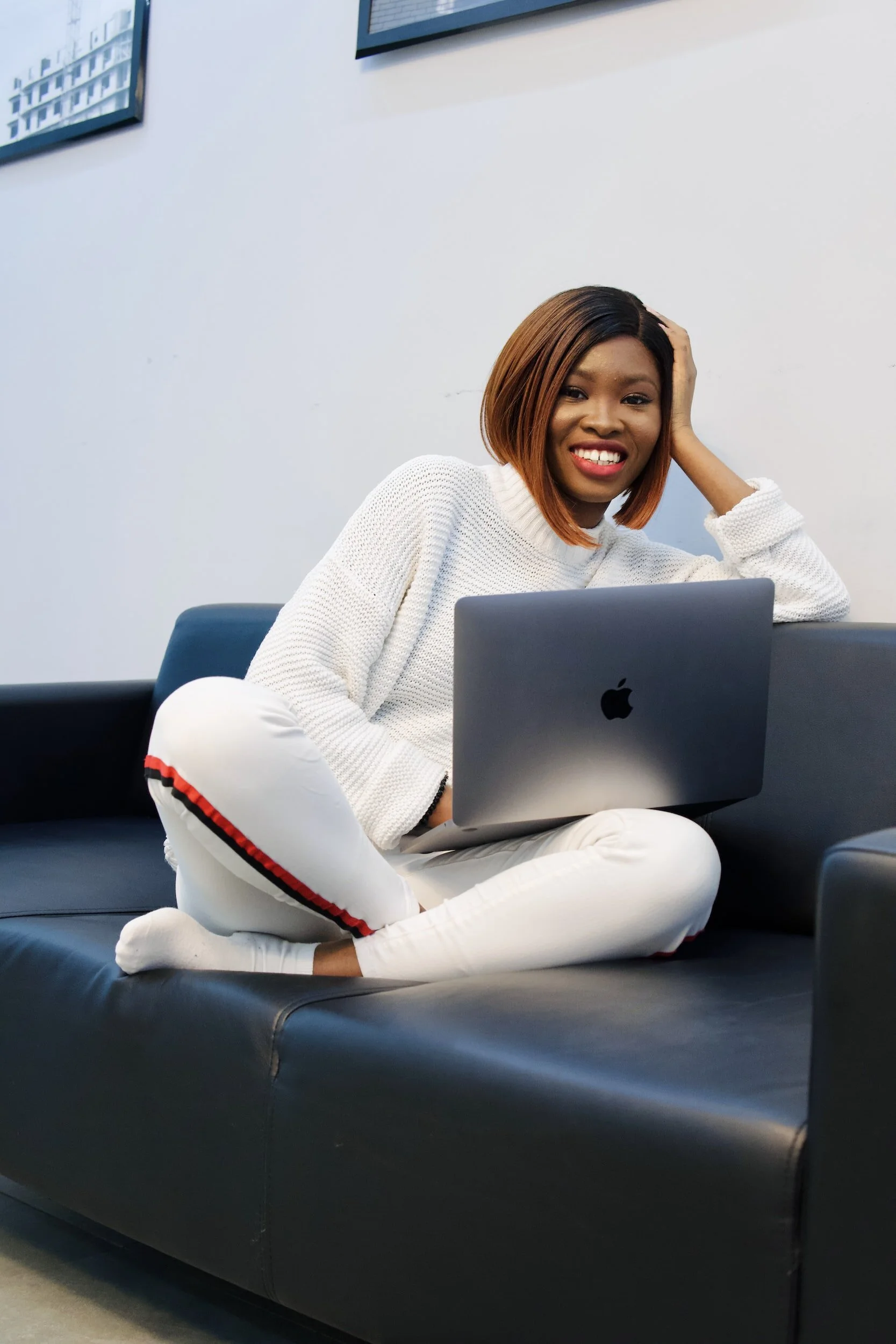 A woman with medium-length brown hair and a white sweater, sitting on a black leather sofa with her legs crossed, smiling at the camera with a silver laptop on her lap, in a room with white walls and framed artwork.