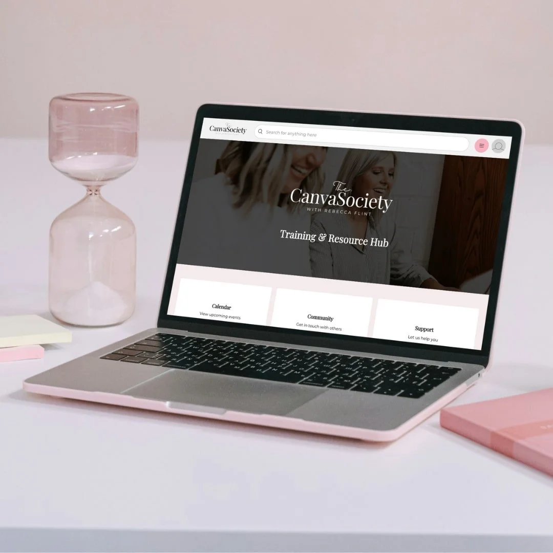 A laptop open on a white desk displaying a website called The Canva Society, with a pink hourglass and pink notebook nearby and a glass of water to the side.