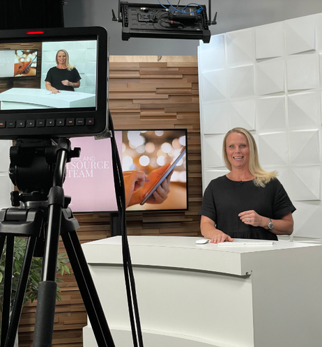 Rebecca Flint,A woman with long blonde hair in a black dress sitting at a white desk in a studio, being filmed from a camera in front of her. The background includes a wood panel wall, a white paneled wall, and two screens displaying related content.