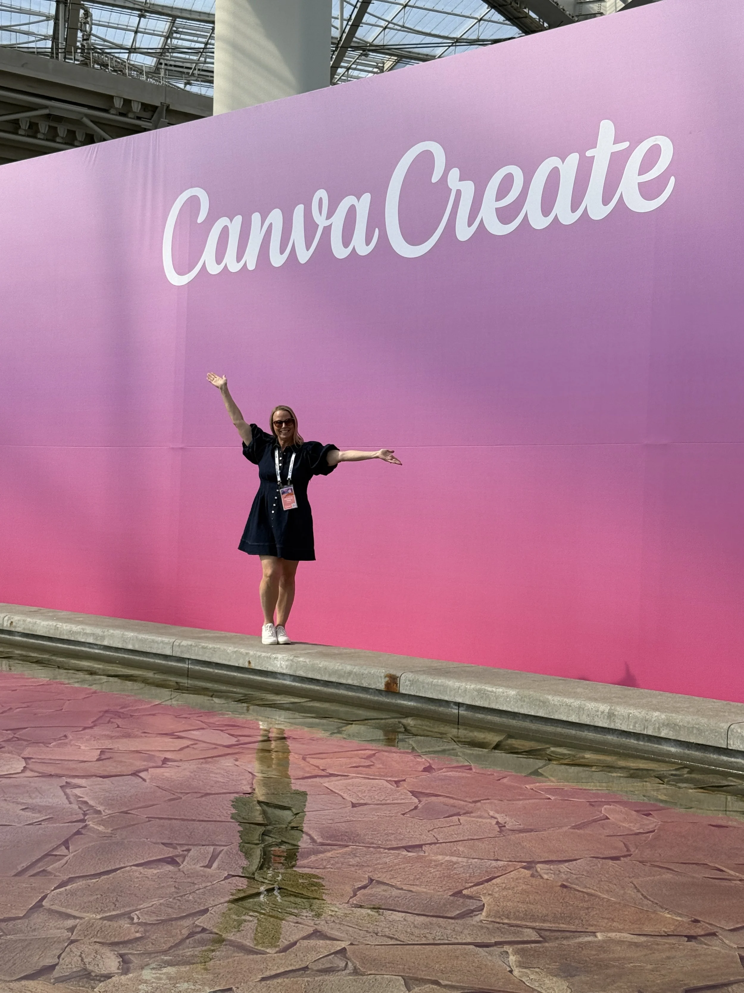 Rebecca Flint, Woman standing with arms outstretched in front of a large pink sign that says "CanvaCreate" in white cursive letters, with a modern building with a glass ceiling reflecting the scene and a puddle of water on the stone floor.