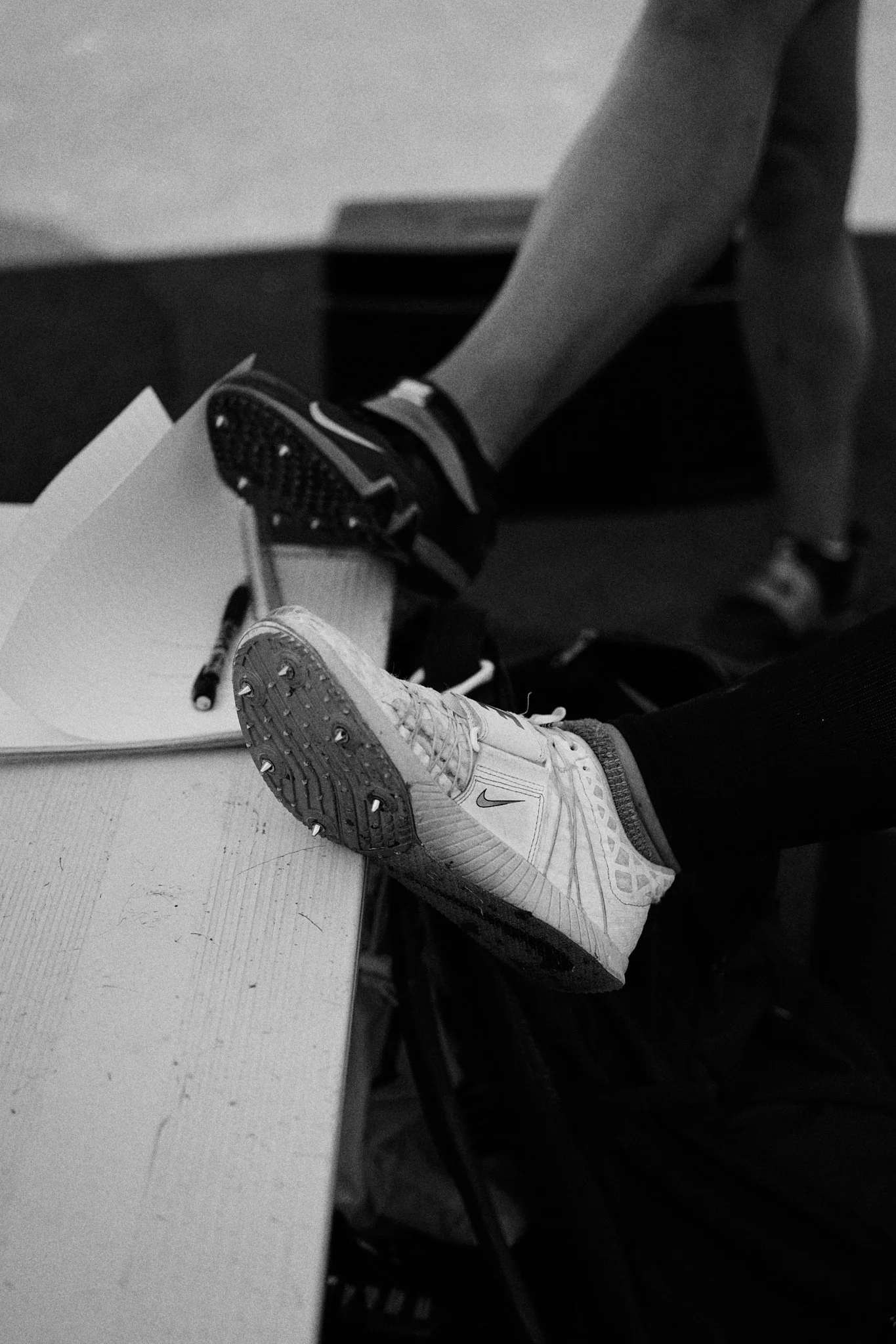 Close-up of a person's sneaker on a table, with another person's foot in a different sneaker hanging over the edge, and papers with a marker nearby.