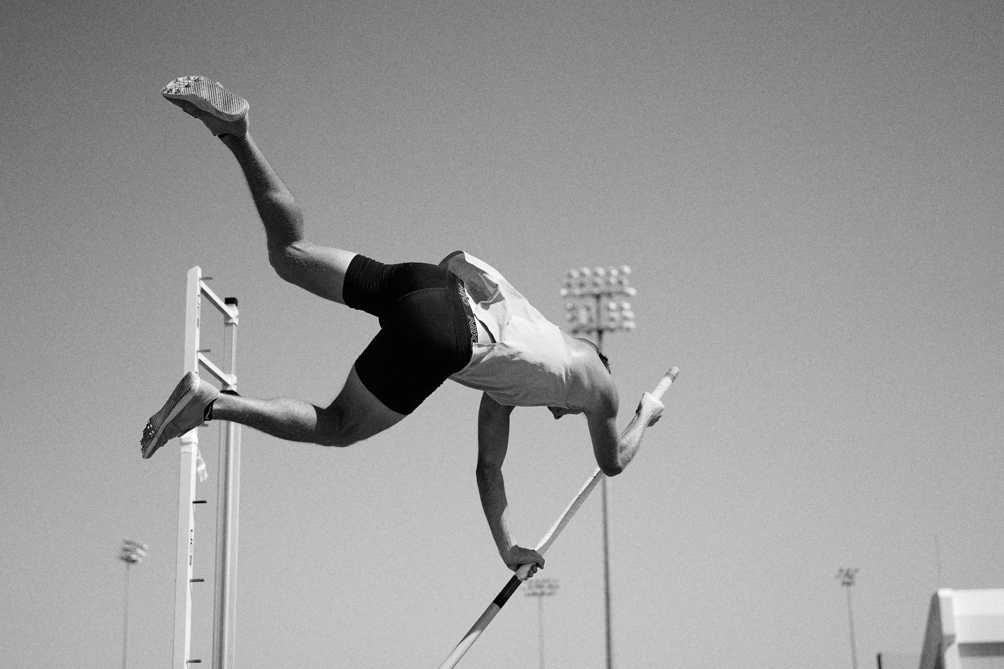 A male pole vaulter in mid-air attempting a vault during a track and field event outdoors in sunny weather.