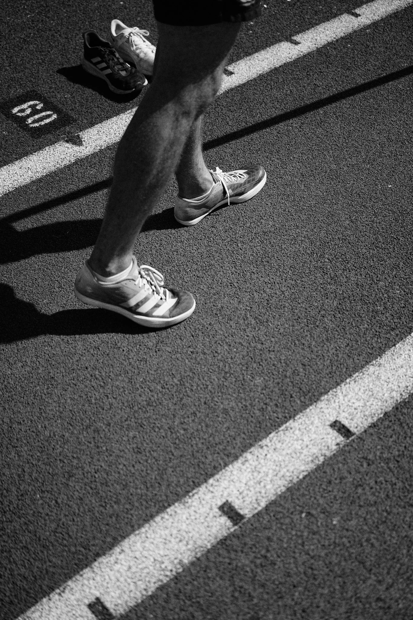 Close-up view of runner's legs and feet on track, wearing sneakers, with another person's shoes in the background, numbered lane markings visible on the track.