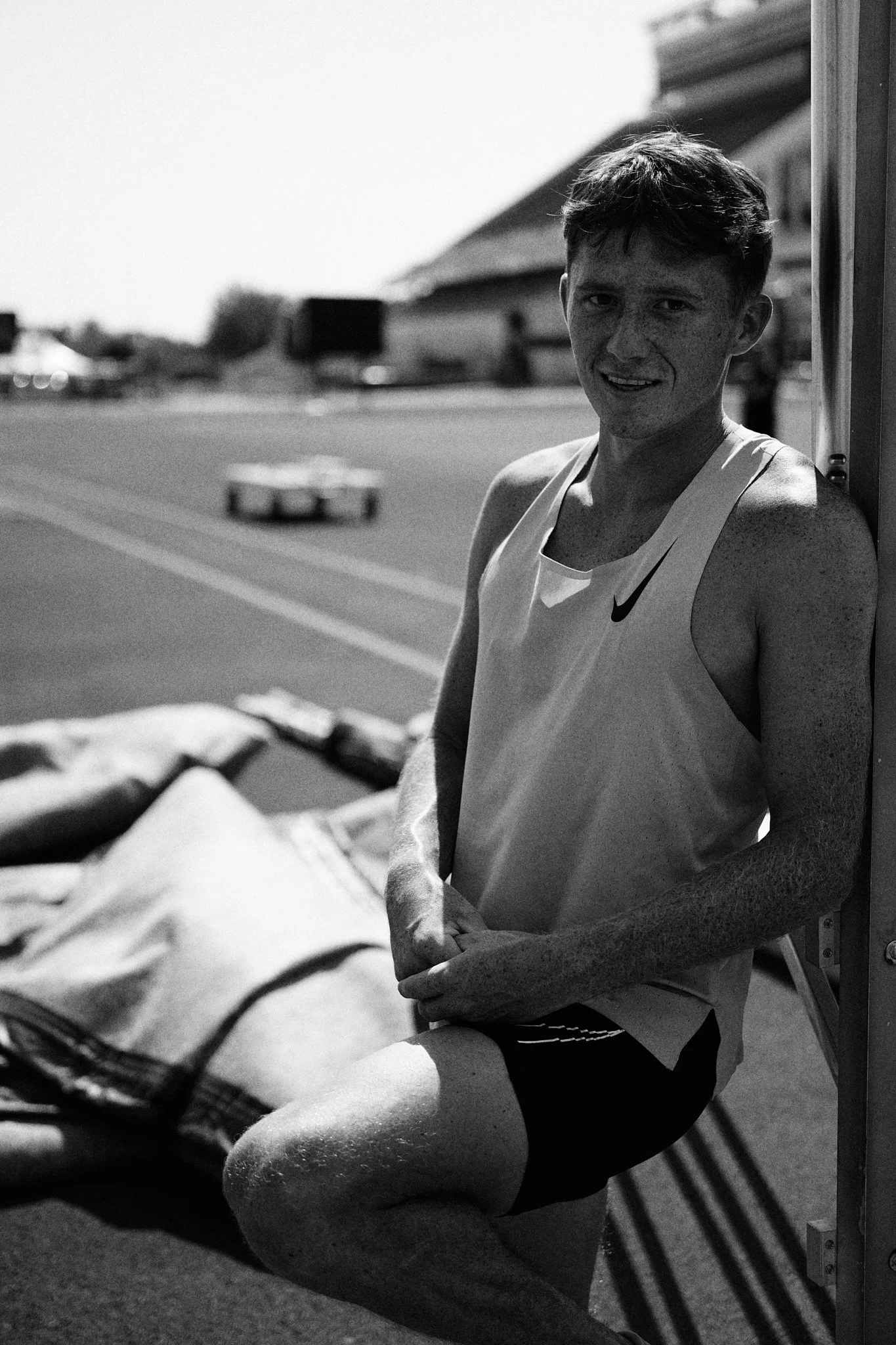 A young man in athletic attire sitting on the ground next to a wall, outdoors on a running track, with sports bags behind him, smiling at the camera in black and white photo.
