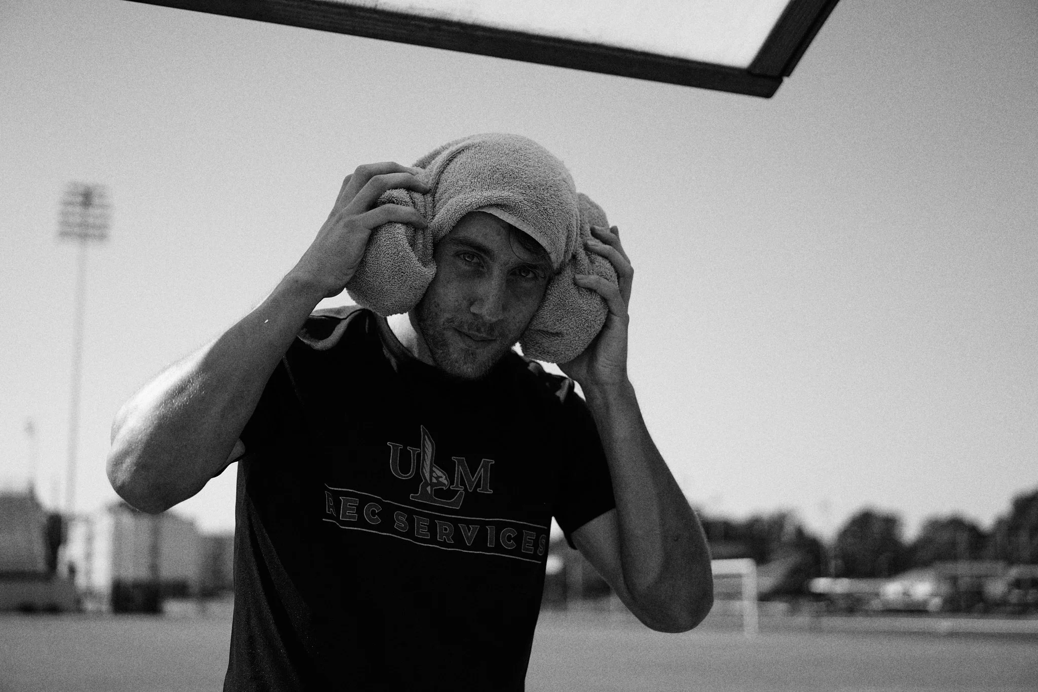 A man wearing a towel over his head and a sports shirt, standing on a soccer field, wiping sweat with a towel and looking at the camera.
