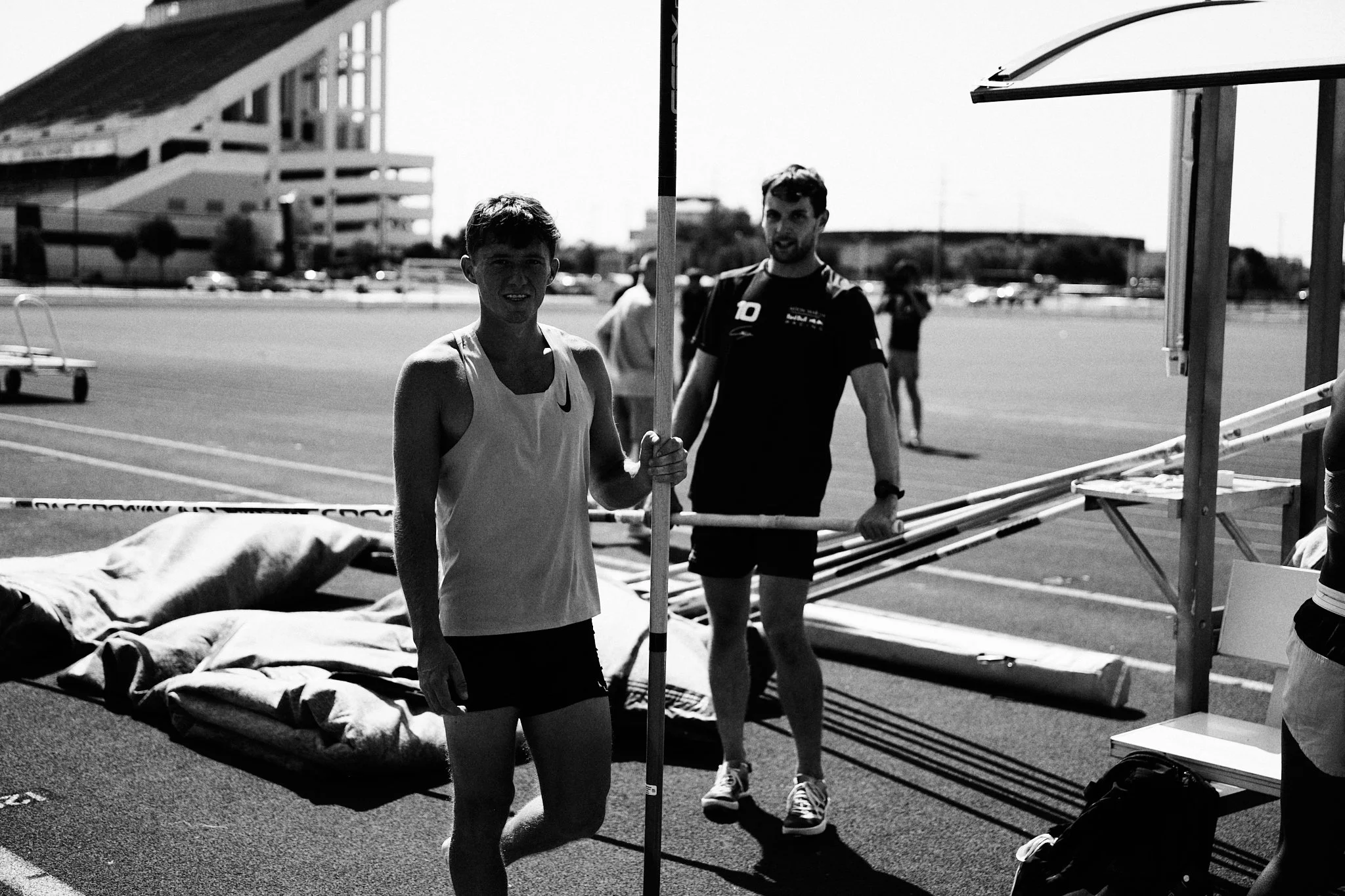 Two male athletes preparing for a pole vault event outdoors, with mats and equipment nearby, on a track field on a sunny day.