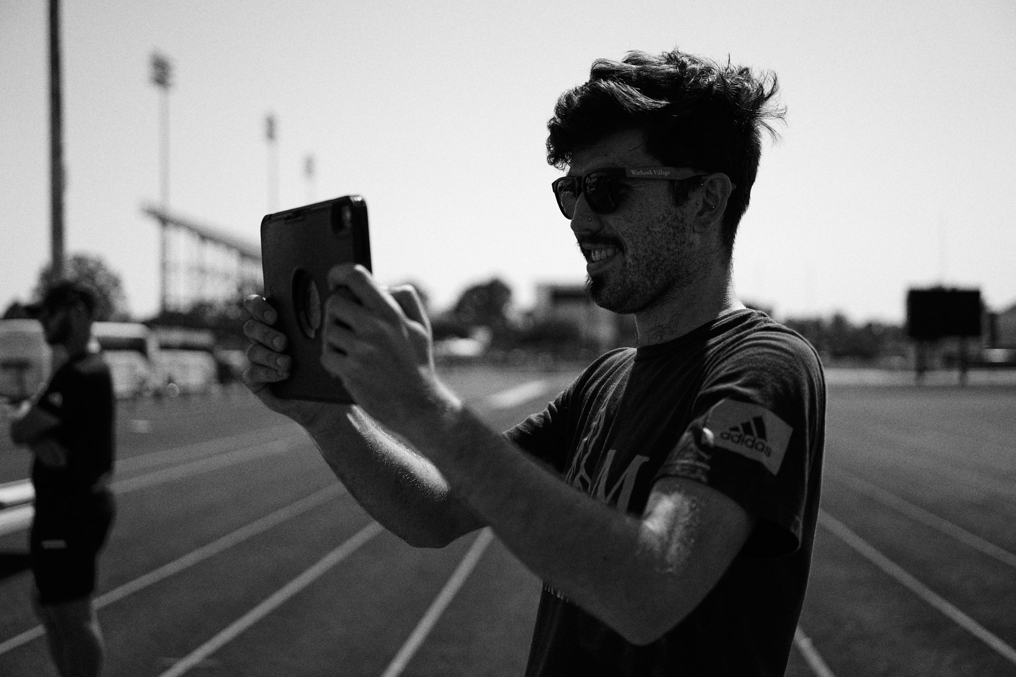 A man with sunglasses and a patterned shirt smiles while taking a photo with his phone at a running track, with another person standing in the background.