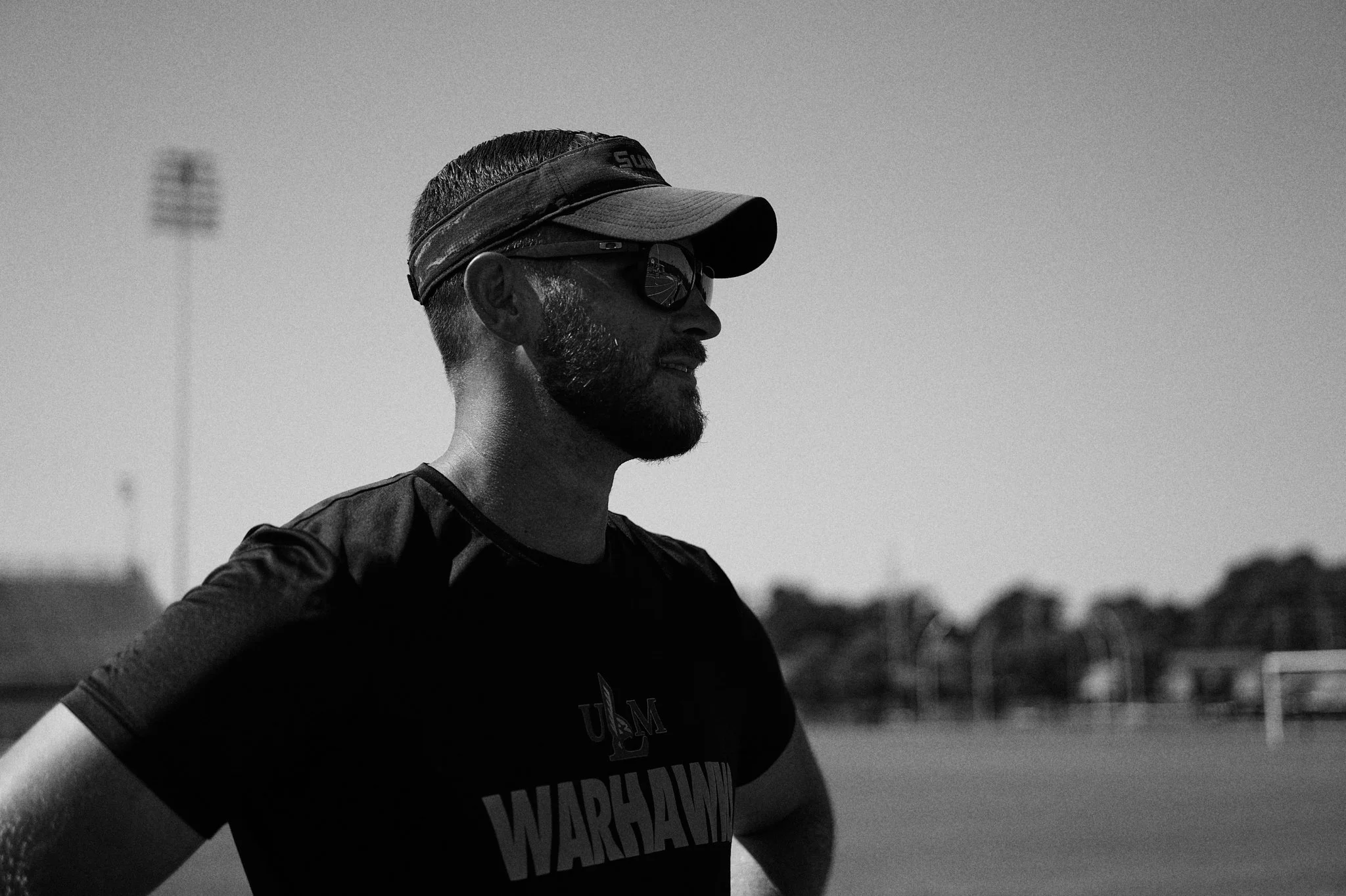 A man with a beard wearing sunglasses, a cap, and a sports shirt standing outdoors on a sports field, with a slightly cloudy sky in the background.
