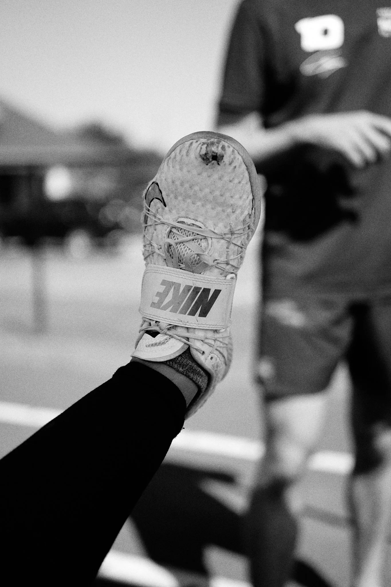 A close-up of a person’s foot wearing a Nike sneaker with a strap, raised in the air during a sports event, with an athlete in the background.