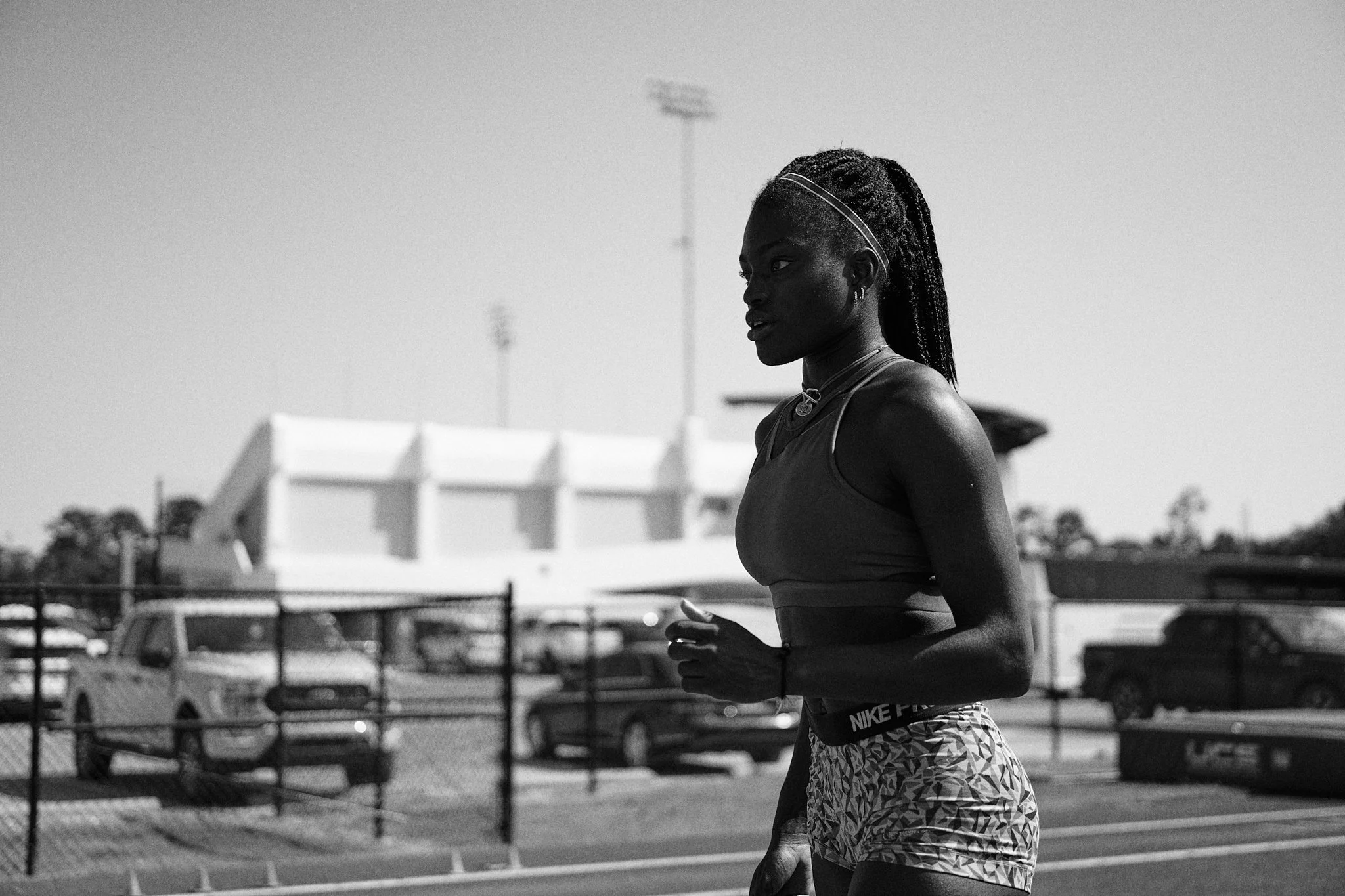A woman running outdoors on a track field with a fence and parked cars in the background, in black and white.