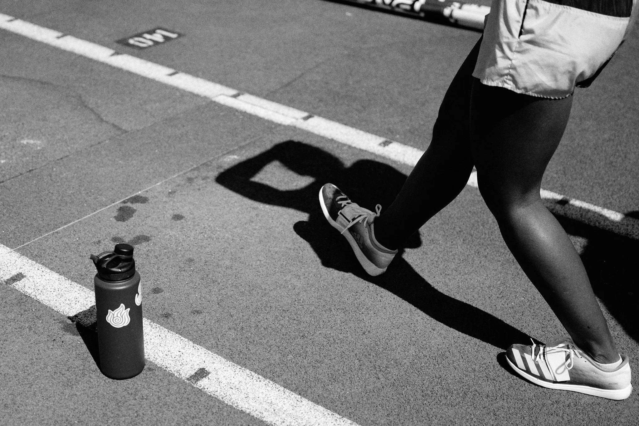 A woman walking outdoors wearing athletic shoes and leggings, with a water bottle on the ground nearby, casting a shadow.