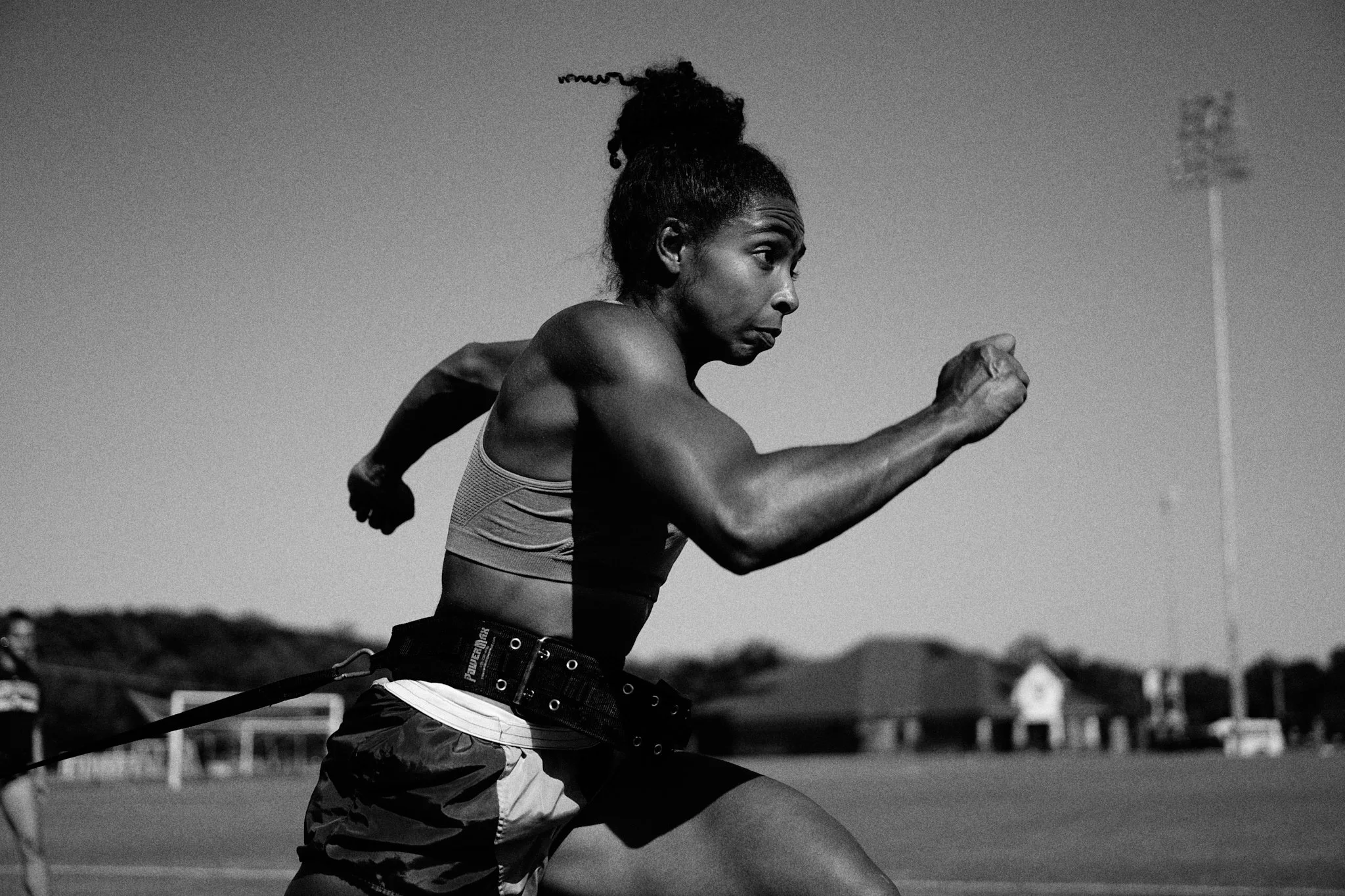 Black and white photo of a female athlete sprinting outdoors on a track, wearing a sports bra and shorts, with her hair tied in a bun, focused on her running.