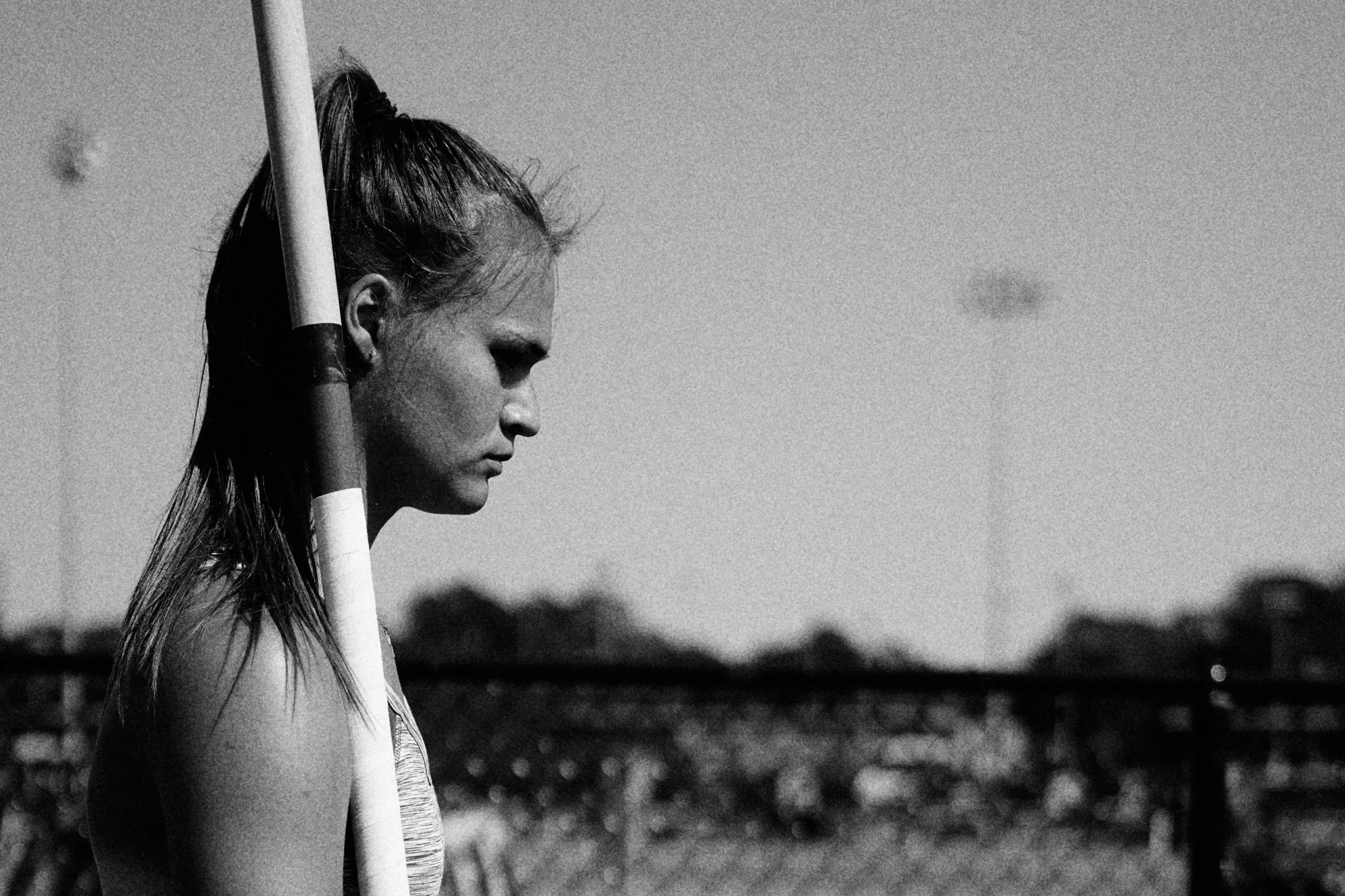 A black and white photo of a woman holding a hockey stick on an outdoor field, with a fence and sky in the background.