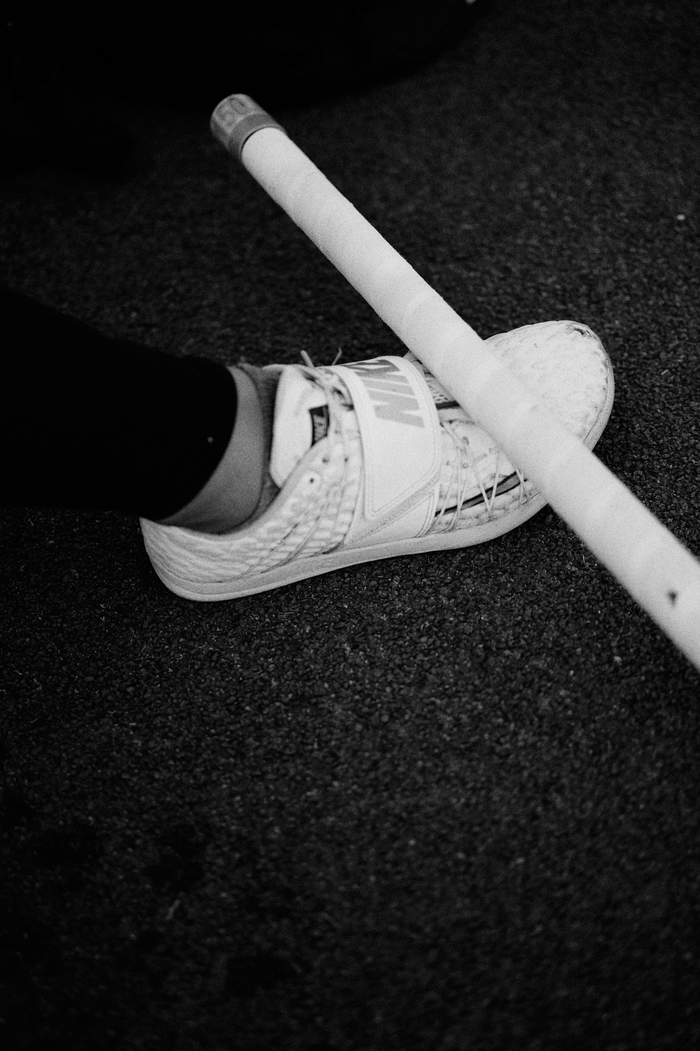 A close-up black and white photo of a person's foot wearing a white sneaker with a black sock, resting on dark textured ground, with a broken hockey stick lying across the shoe.