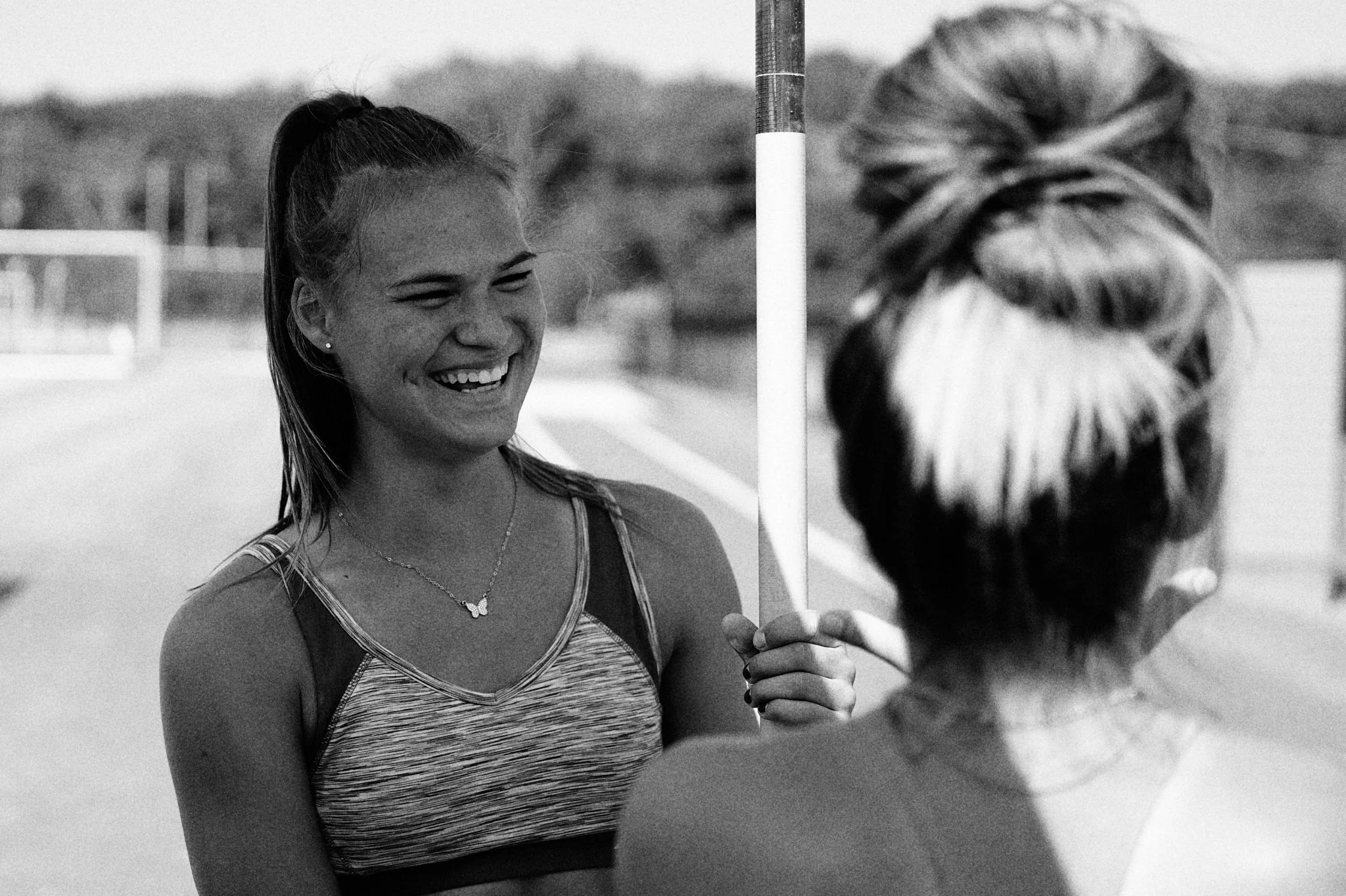 Two young women at an outdoor track field, one smiling and holding a pole vault pole, the other with back to the camera.