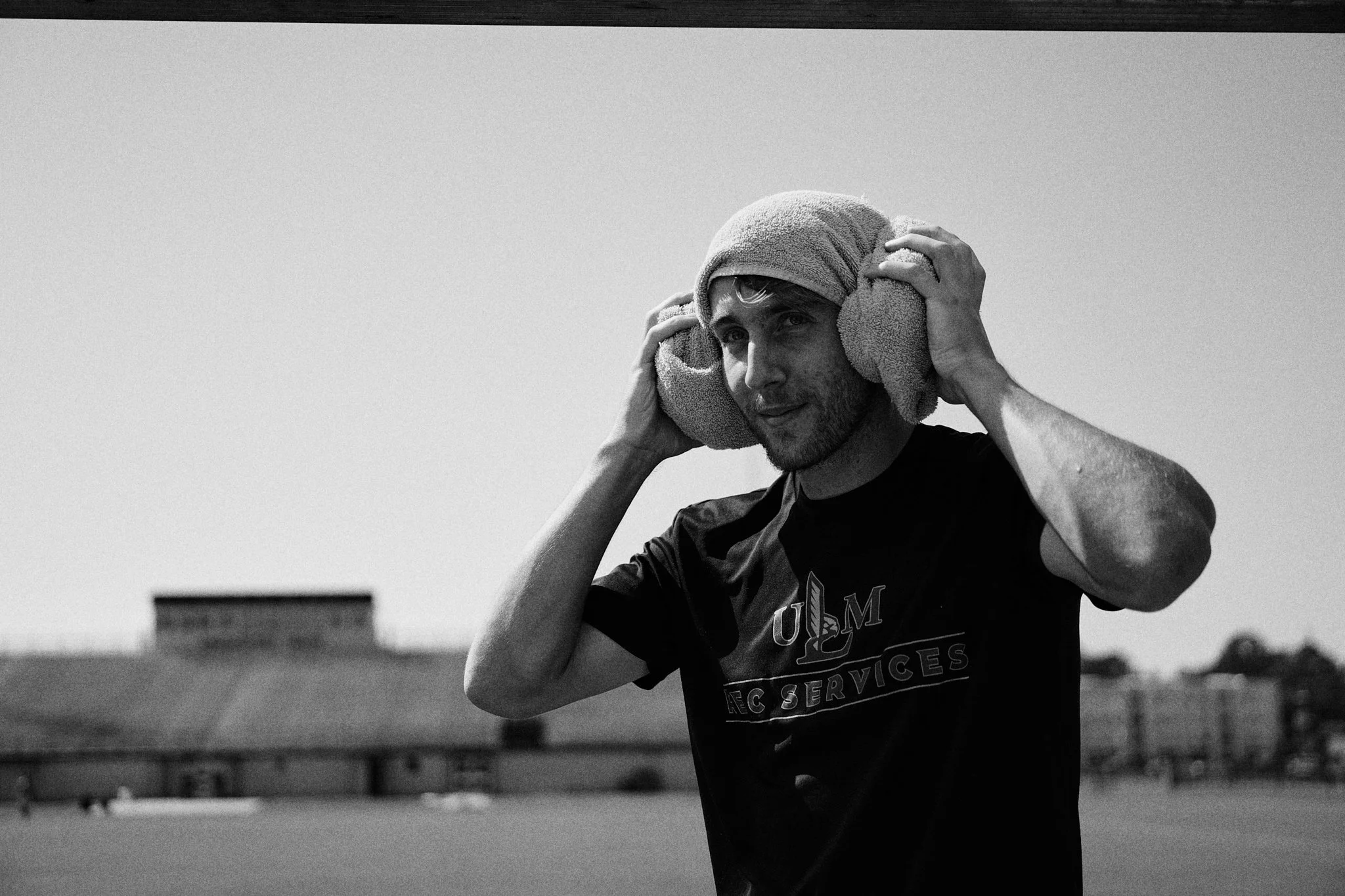 A man wearing a black shirt and a towel on his head is holding headphones to his ears in an outdoor setting, with industrial buildings in the background.